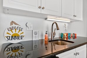 A kitchen with white cabinets, a stainless steel sink, and decorative wall stickers. - Endless Summe, Oceanfront 8 Bedroom w/ Heated Pool (Holden Beach)