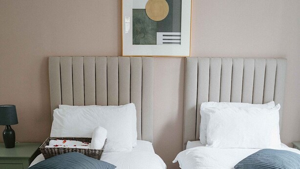 Une chambre d’hôtel avec deux lits, chacun doté d’une tête de lit, de draps blancs et d’oreillers bleus. Un tableau encadré est accroché au-dessus des lits, et une table de chevet avec une lampe se trouve à gauche.