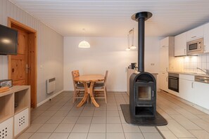 A compact kitchen with a wood-burning stove, a dining table with chairs, a TV mounted on the wall, and a microwave. - Gemutlicher Doppelbungalow mit Garten und Terrasse im Ferienpark Mirow (Mirow)