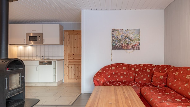 Une cuisine moderne avec des meubles blancs, un four à micro-ondes et un four encastré. Le salon est équipé d’un banc en bois et d’un canapé rouge à motifs.