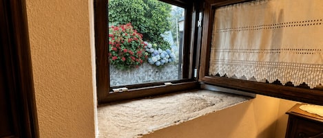 A room with a window overlooking a garden with red and blue flowers, a wooden bench, and a wooden dresser.