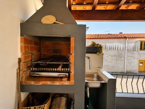 An outdoor kitchen with a brick oven, a sink, and a window overlooking a tiled roof. - Casa dos Laia | Nature, Comfort, and Tranquility (Sobreira Formosa)