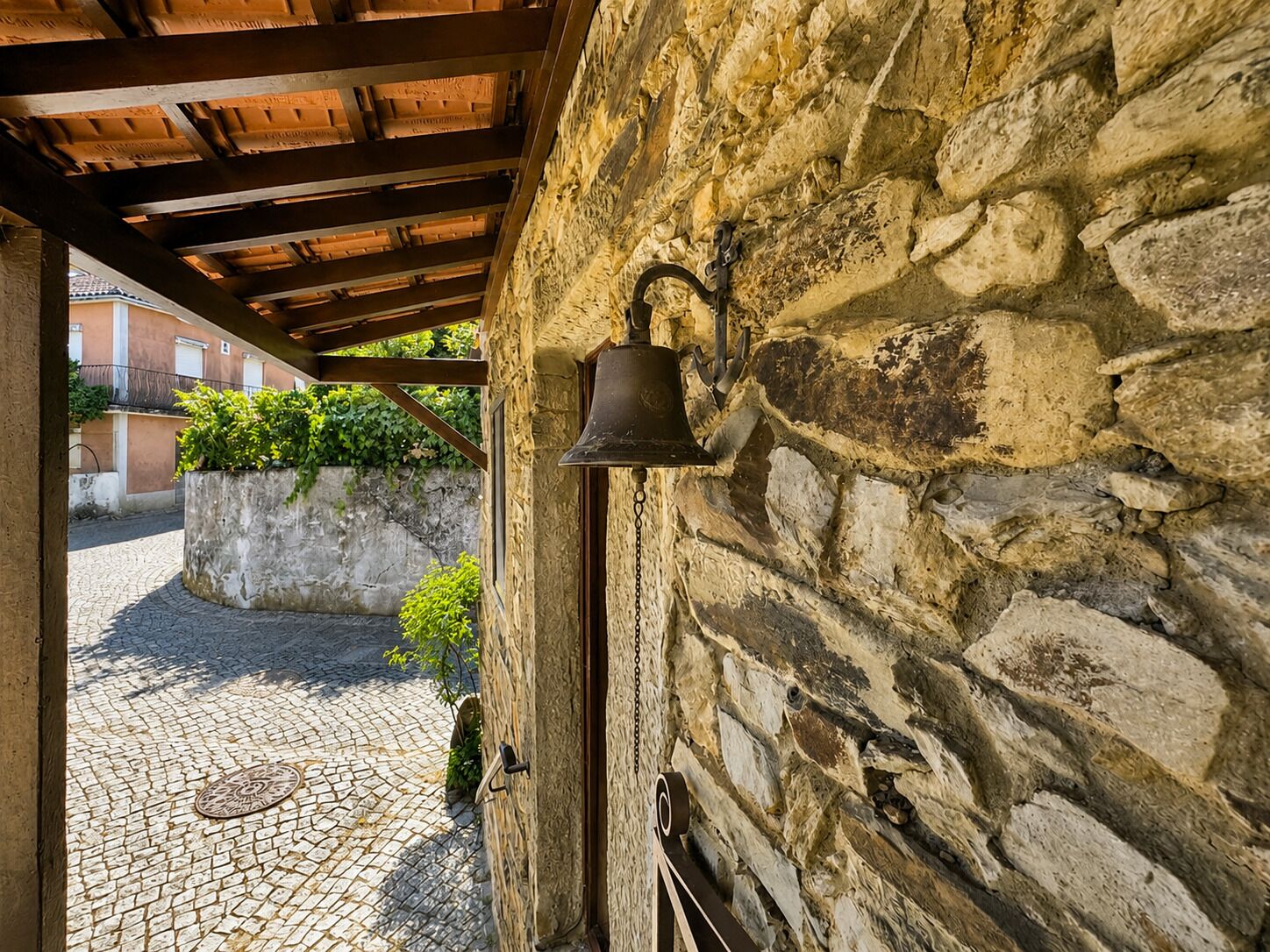Um muro de pedra com um sino e uma porta, uma rua de paralelepípedos e um edifício ao fundo.