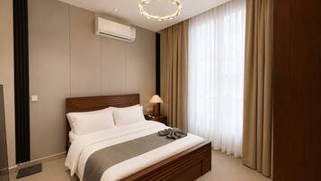 Una habitación de hotel con cama, unidad de aire acondicionado, mesita de noche, lámpara y un ventanal con cortinas.