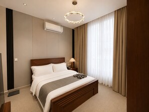 Una habitación de hotel con cama, unidad de aire acondicionado, mesita de noche, lámpara y un ventanal con cortinas.