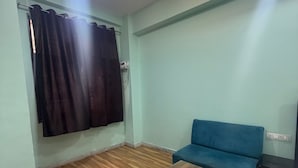 A room with a wooden floor, a blue sofa, a small wooden table, a ceiling light, and a window with a curtain. - A.V Grand Hotel (Kanpur)