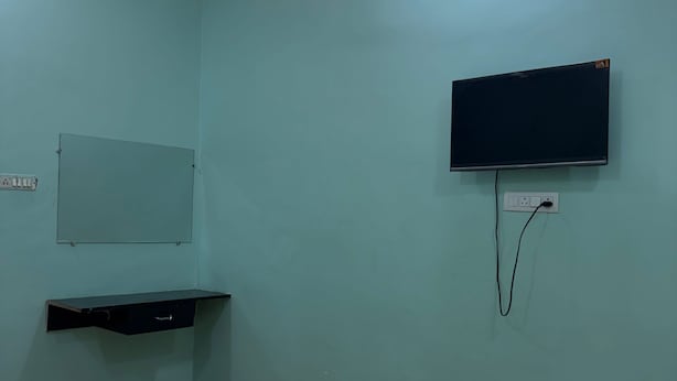 A room with a bed, a flat-screen TV mounted on the wall, a power outlet, and a shelf. - A.V Grand Hotel (Kanpur)