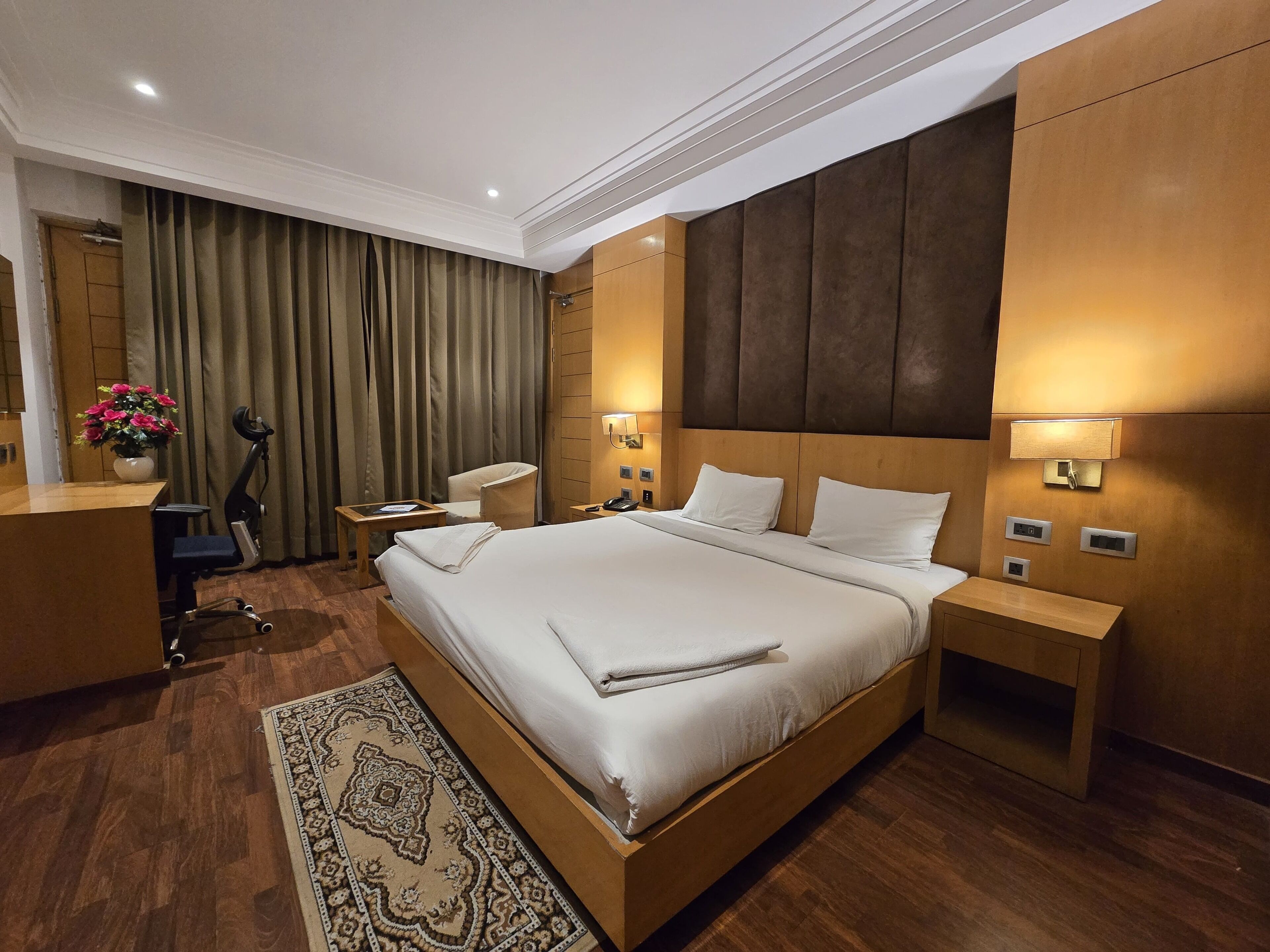 A hotel room with a large bed, a desk, a chair, a desk lamp, a nightstand, a bedside table, and a wall-mounted lamp.