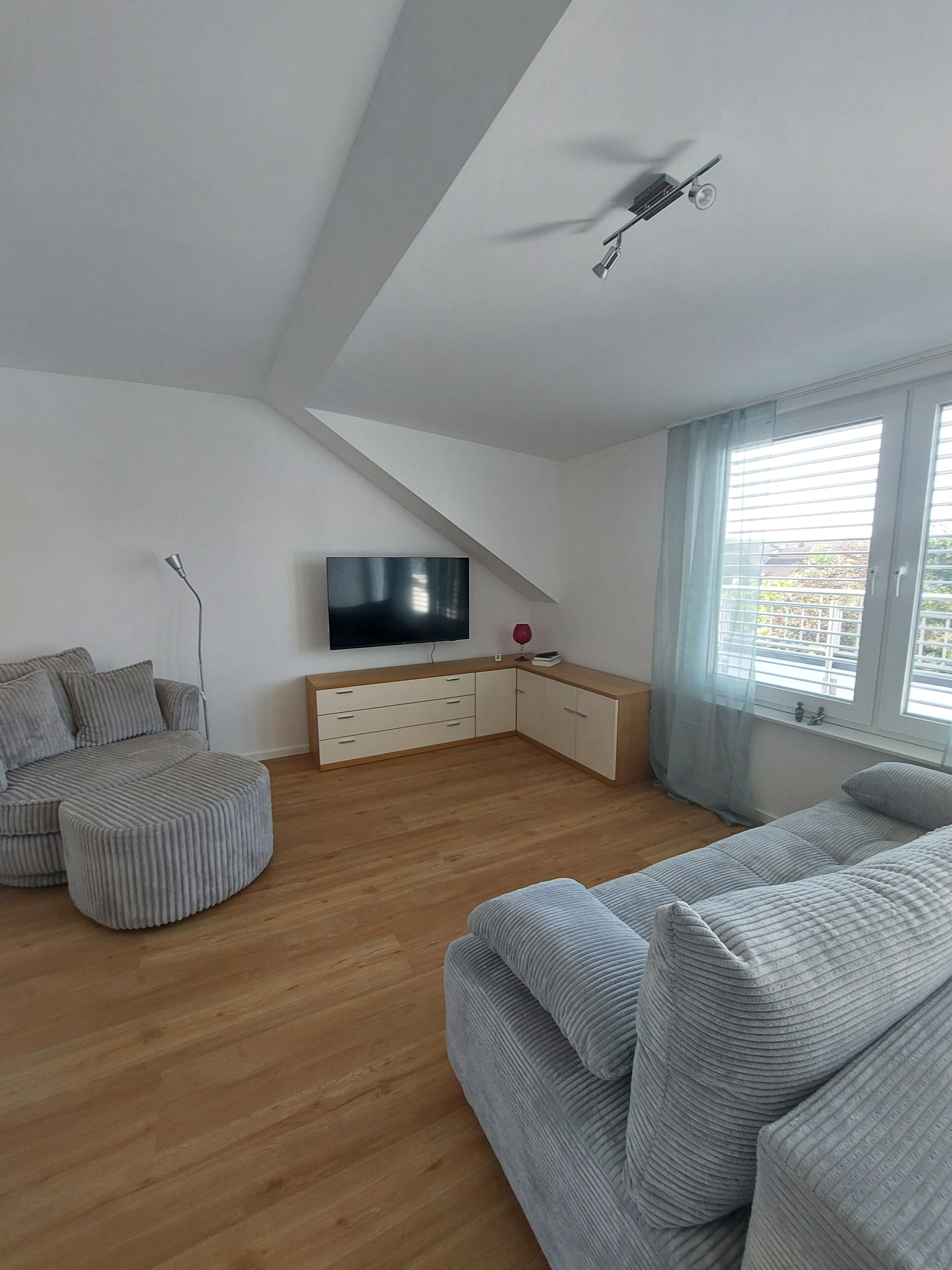 Un salon moderne avec un plafond en pente, un téléviseur à écran plat, un meuble en bois et une grande fenêtre équipée de stores.