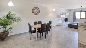 A modern kitchen with a dining area, a clock on the wall, and a large window providing natural light. - Fully equipped 3BR Mosta home, Rotunda View w/WIFI (Mosta)