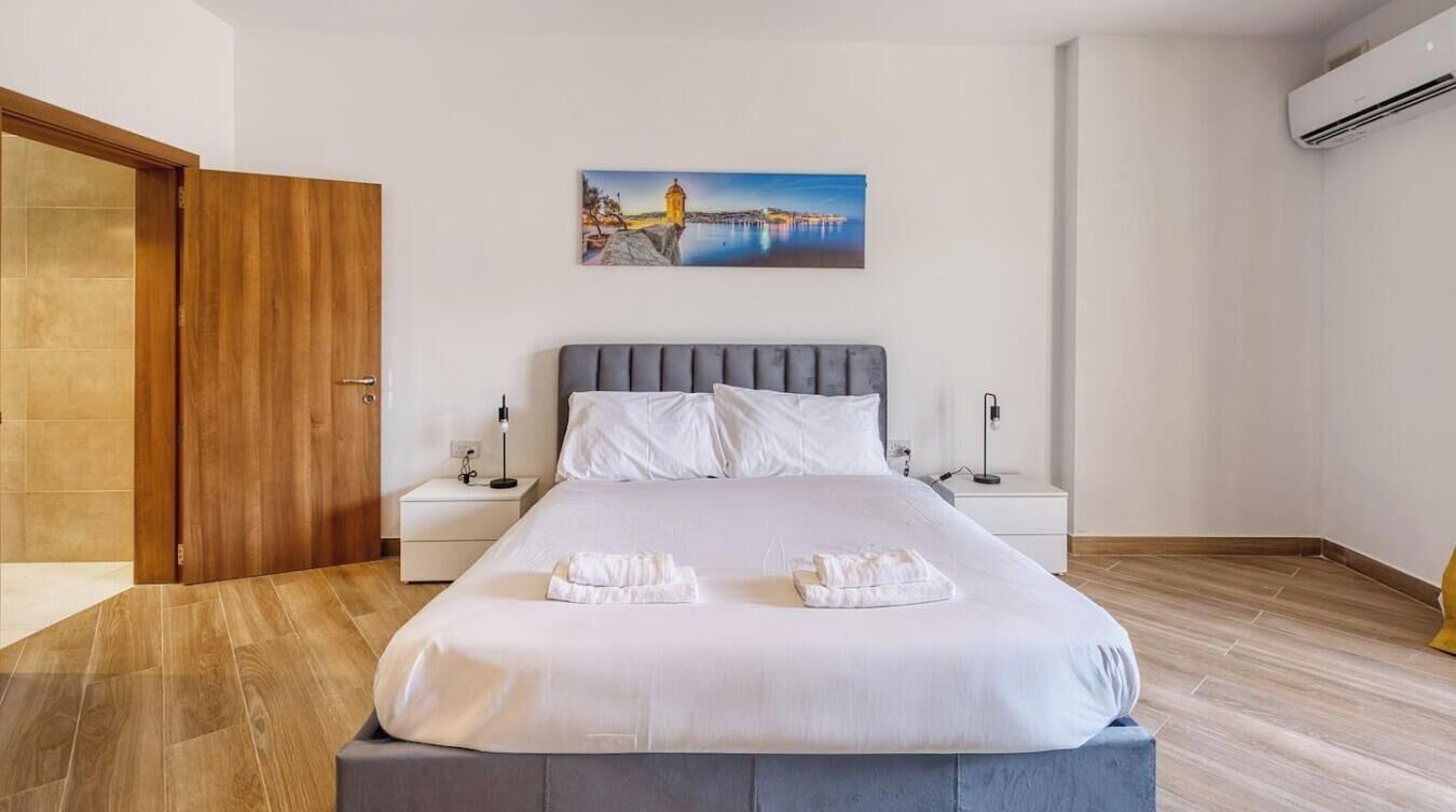Hotellihuone, jossa on sänky, kaksi yöpöytää, yölamppu, taulu seinällä ja ilmastointilaite.