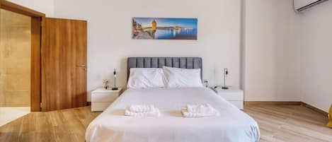A hotel room with a bed, two bedside tables, a nightstand, a painting on the wall, and an air conditioning unit.