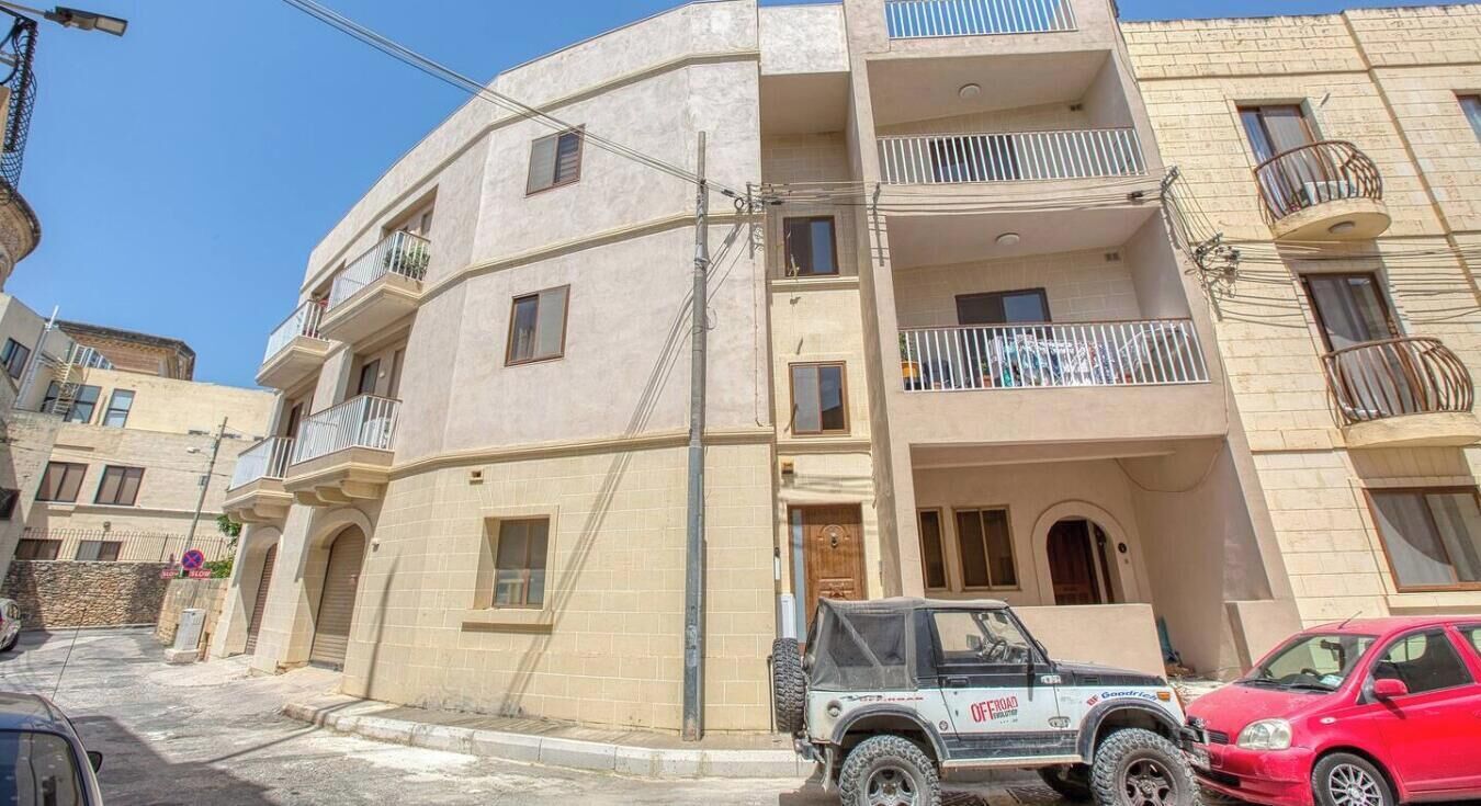Un edificio de varios pisos con balcones, un vehículo todoterreno estacionado y un coche rojo.