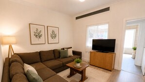 A living room with a sectional sofa, a coffee table with a plant, a television on a wooden stand, and framed botanical prints on the wall. - Generous 4BDR Modern Family Home in Ilford Lane (Greater London)