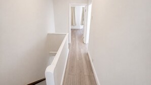 A narrow hallway with a wooden floor, white walls, and a door at the end leading to a room with a shower. - Generous 4BDR Modern Family Home in Ilford Lane (Greater London)
