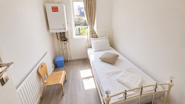 A small, single-bed room with a radiator, a window with curtains, a chair, and a blue trash bin. - Generous 4BDR Modern Family Home in Ilford Lane (Greater London)