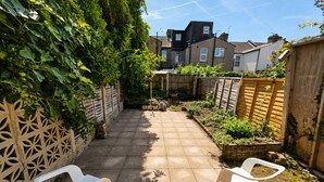 A paved patio with two white plastic chairs, a wooden fence, and a garden area with plants and a brick wall. - Generous 4BDR Modern Family Home in Ilford Lane (Greater London)