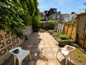 A paved patio with two white plastic chairs, a wooden fence, and a garden area with plants and a brick wall. - Generous 4BDR Modern Family Home in Ilford Lane (Greater London)