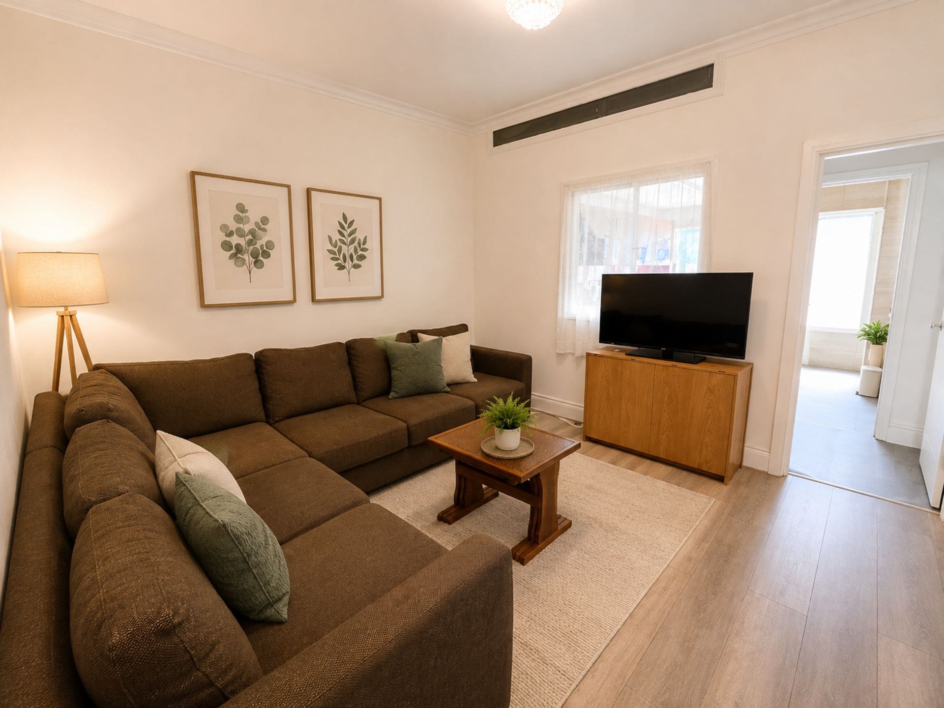 Ein Wohnzimmer mit einem Ecksofa, einem Couchtisch mit Pflanze, einem Fernseher auf einem Holzboden und gerahmten botanischen Drucken an der Wand.