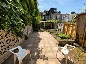 A paved patio with two white plastic chairs, a wooden fence, and a garden area with plants and a brick wall. - Generous 4BDR Modern Family Home in Ilford Lane (Greater London)