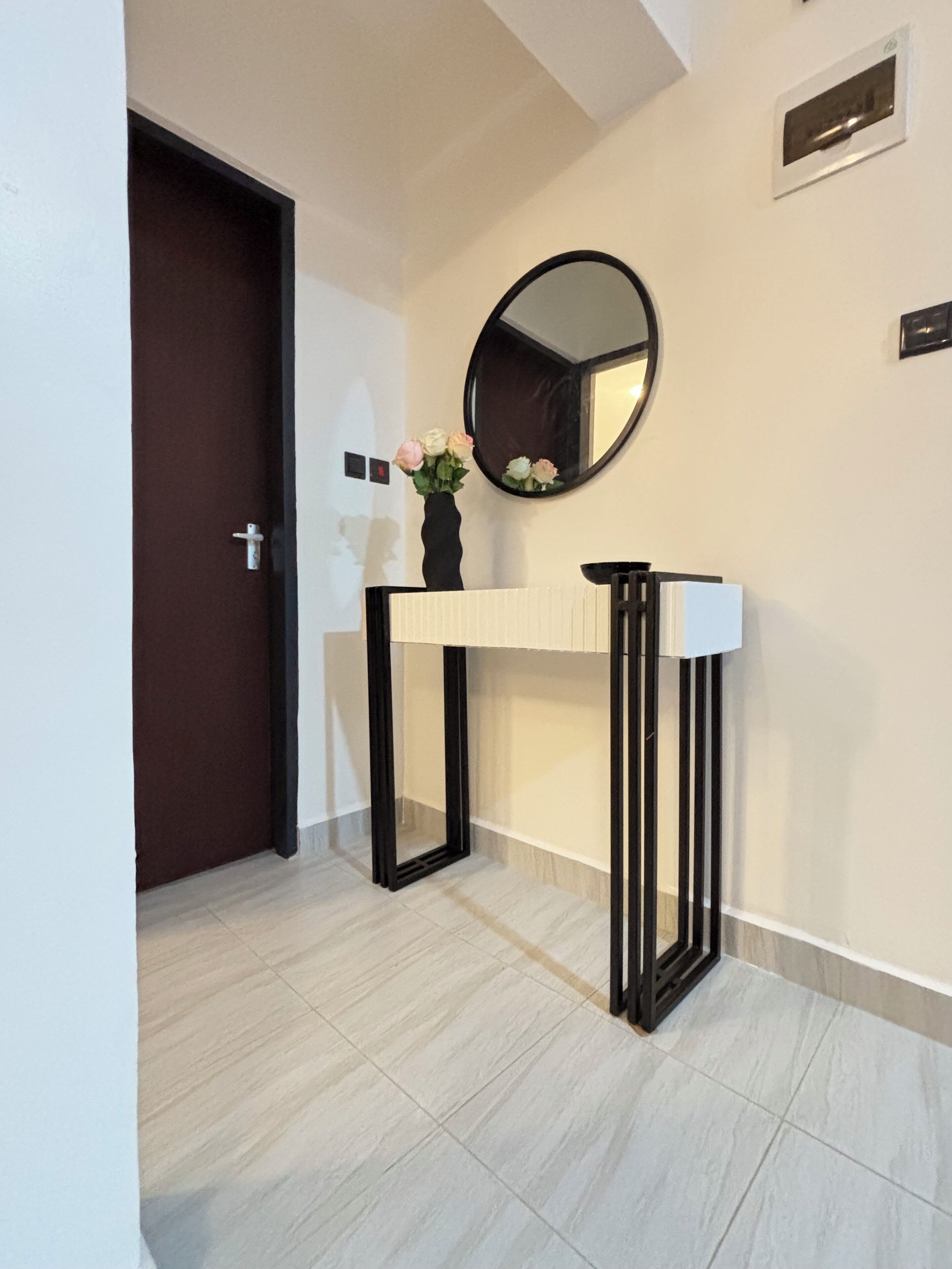 Un corridoio con una porta, un tavolo consolle con un vaso di fiori, uno specchio rotondo e un interruttore a parete.