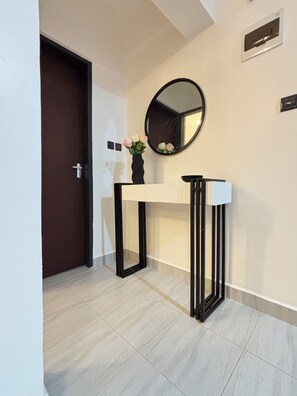 A hallway with a door, a console table with a vase of flowers, a round mirror, and a light switch on the wall. - Luxury 1BR Near Two Rivers & Karura Fast Wi-Fi Free Parking Lift Backup Power (Kiambu County)