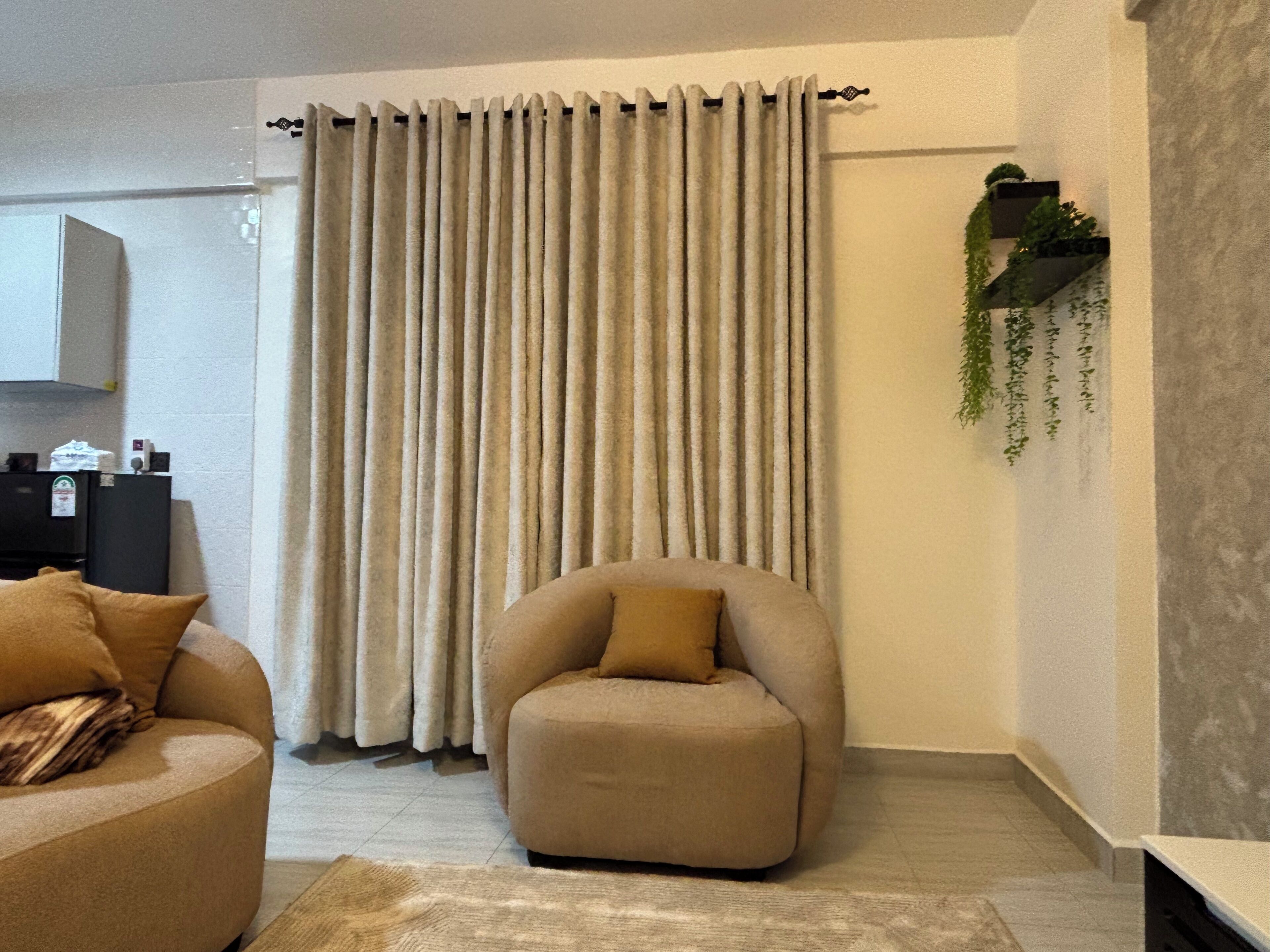 Un salon avec un canapé beige, un fauteuil beige, une grande fenêtre avec des rideaux beiges, une petite plante accrochée au mur et un meuble noir sur lequel sont posés divers objets.