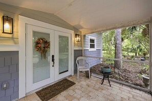 A covered porch with a white door, a glass door, a bench, a potted plant, and a view of the surrounding greenery.