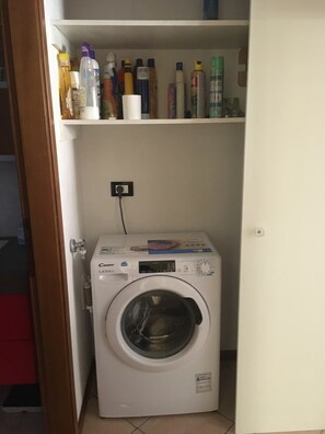 A compact washing machine in a laundry room with a shelf above containing various bottles.