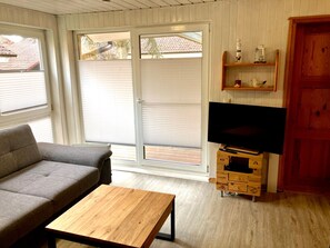 Una sala de estar con sofá, mesa de centro, televisor sobre un mueble de madera y puerta corrediza de vidrio que da a un área exterior.