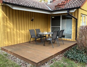 A wooden deck with a table and chairs, a window with blinds, and a yellow wall. - Urlaub am Wasser - Doppelbungalow mit Terrasse im Ferienpark Mirow (Mirow)