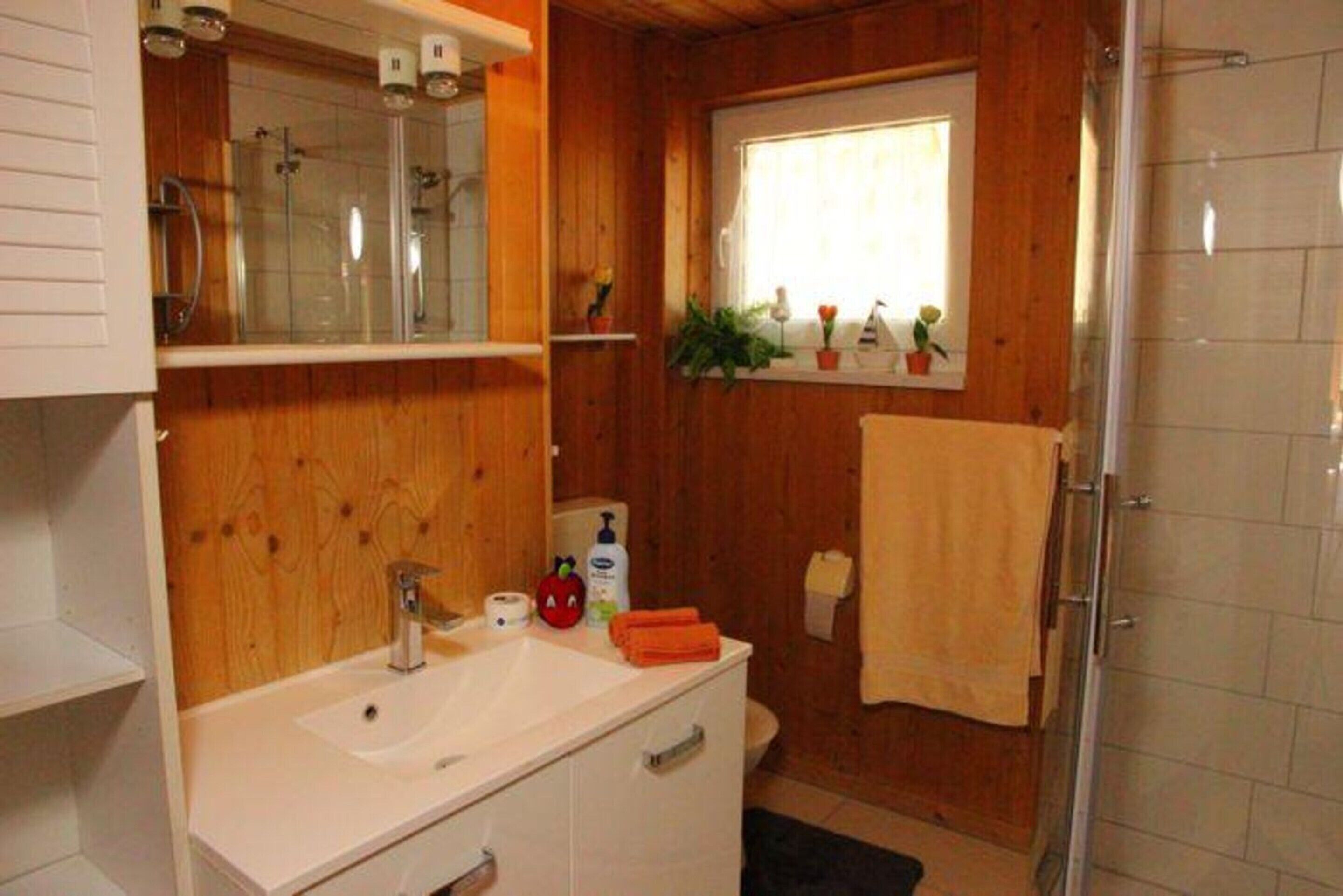 Un baño con paredes de madera, un lavamanos blanco, un espejo, una ducha y una ventana con plantas.