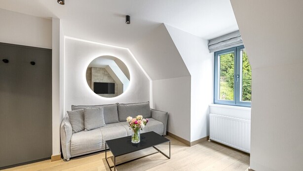 A modern living room with a grey sofa, a round mirror, a black coffee table, and a window with a view of greenery. - Waterfall View Penthouse · One-Bedroom (Residenz Boutique Suites (Bad Gastein))