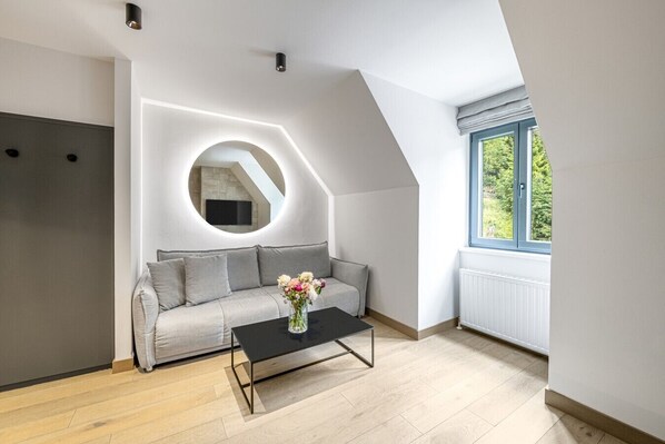 A modern living room with a grey sofa, a round mirror, a black coffee table, and a window with a view of greenery.