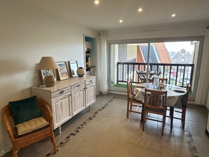 A dining area with a table set for two, a wicker chair, a cabinet with decorative items, and a view of the outside through a large window. - Apartment with balcony, sea view - 4 guests (Cabourg)