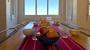 Een eettafel met een fruitschaal, glazen en een karaf, voor een raam met uitzicht op de lucht.