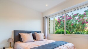 Ein Schlafzimmer mit einem Bett, einem Nachttisch, einer Lampe und einem Fenster mit Blick auf rosa Blumen.
