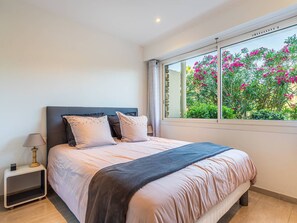 Ein Schlafzimmer mit einem Bett, einem Nachttisch, einer Lampe und einem Fenster mit Blick auf rosa Blumen.