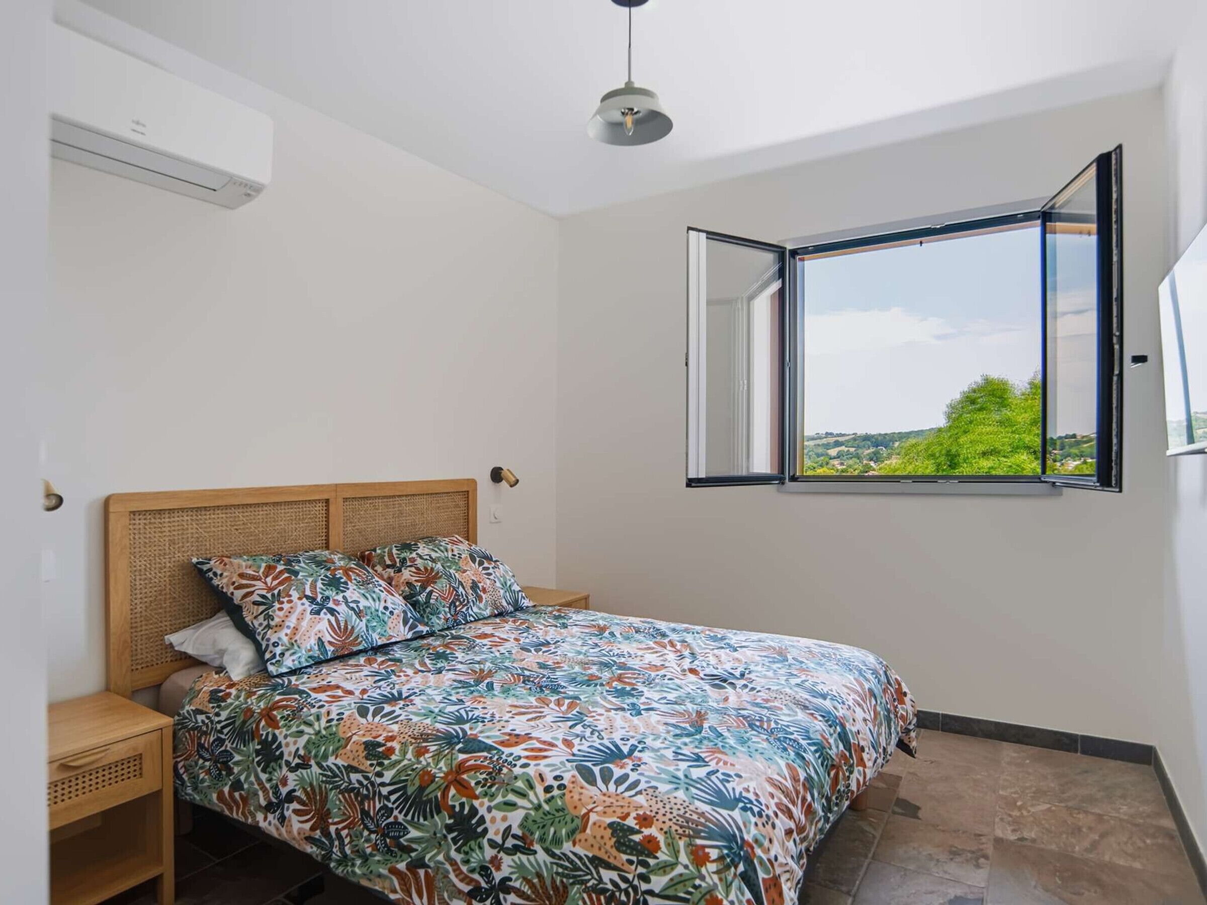 A bedroom with a bed, a window with an open view, a ceiling fan, and a wall-mounted air conditioner.