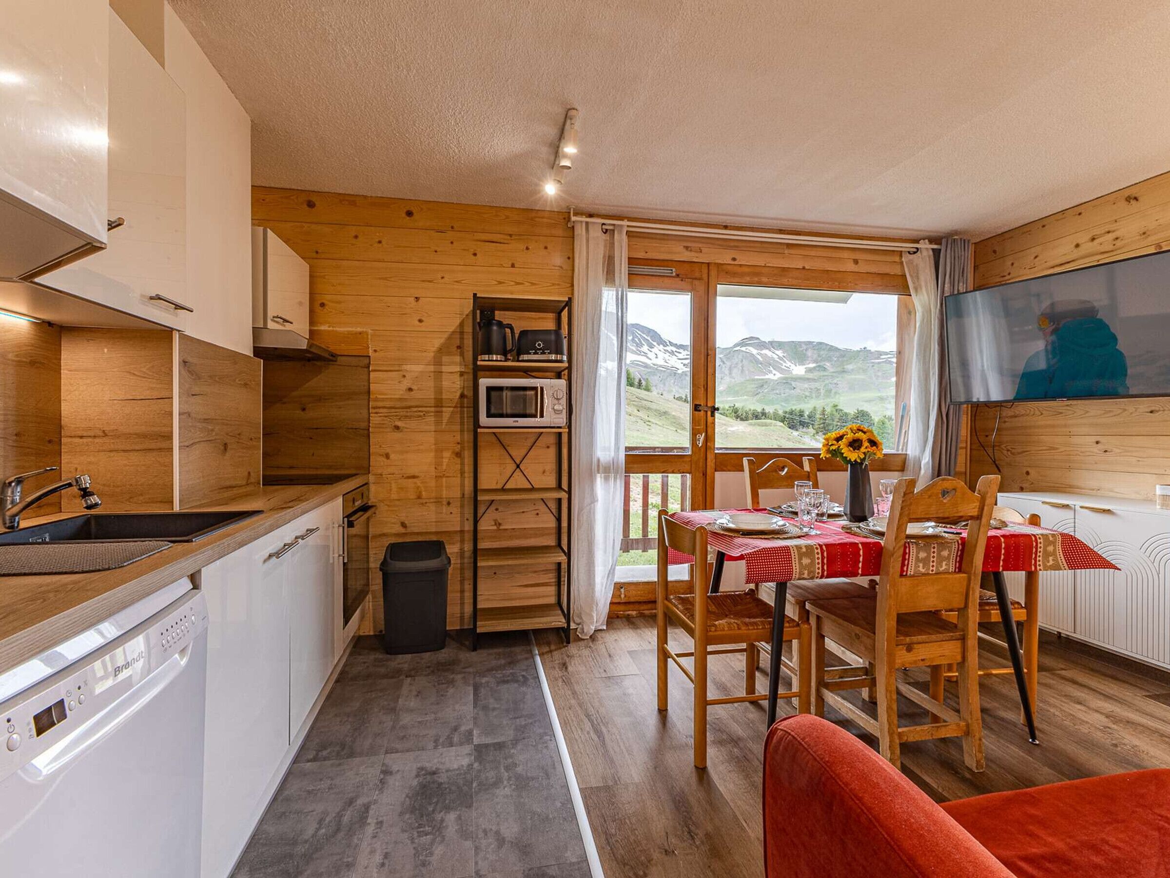 Une cuisine compacte équipée d’un lave-vaisselle, d’un four à micro-ondes et d’un évier. On y trouve également un coin repas avec une table pour deux personnes, un vase de fleurs et une vue sur les montagnes par la fenêtre.