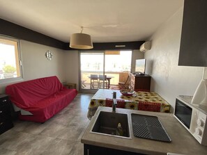 A compact living space with a kitchenette, a red sofa, a dining table with a sunflowers tablecloth, and a view through a sliding glass door. - 2-room apartment, air-conditioned, by the sea for 4 (Canet-Plage)