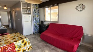 Een knus woongedeelte met een kitchenette, een eethoek met tafel en stoelen, een rode bank en een raam voor natuurlijk licht.