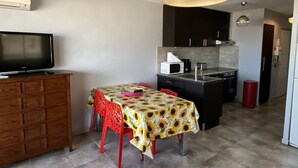 Een ruimtelijk ingerichte leefruimte met een televisie, een eettafel met rode stoelen, een keuken met moderne apparatuur en een grote hanglamp.