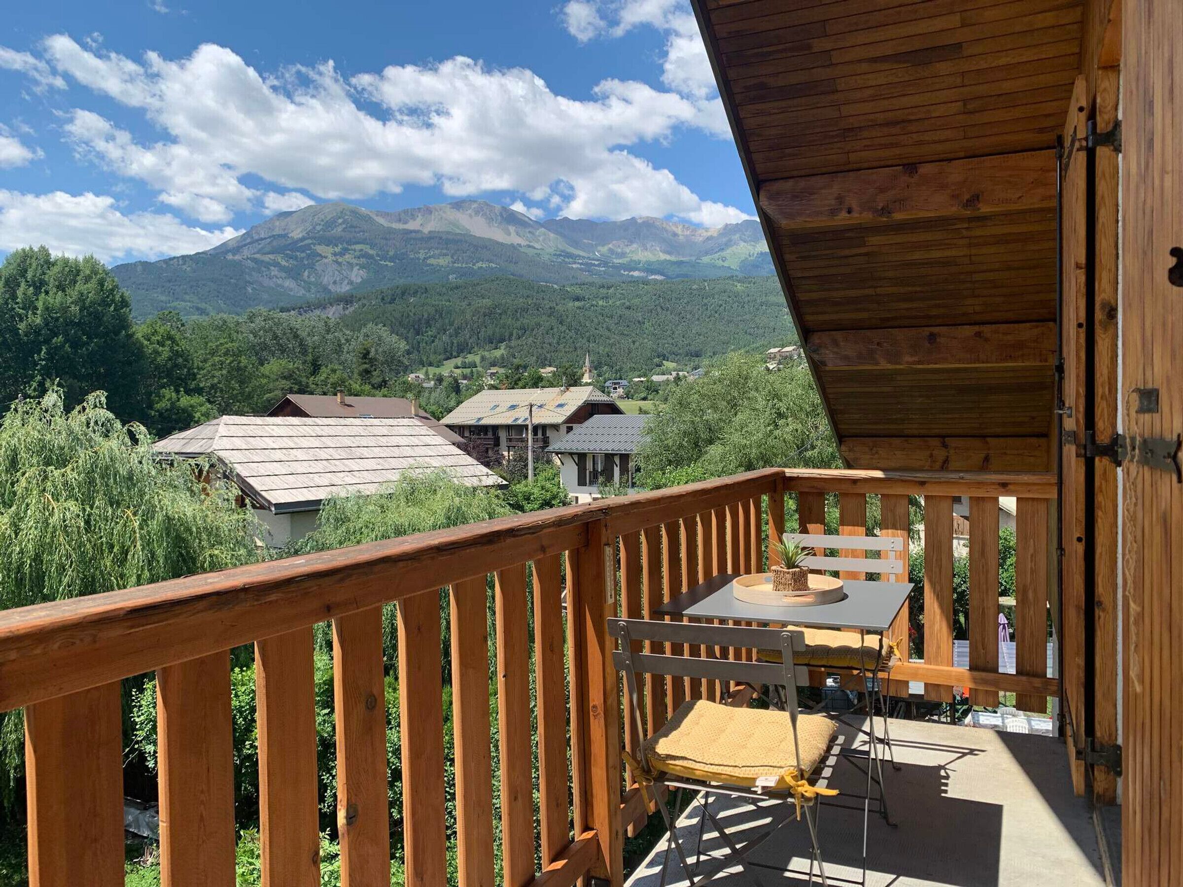 A wooden balcony with a table and chairs, overlooking a mountainous landscape with houses.