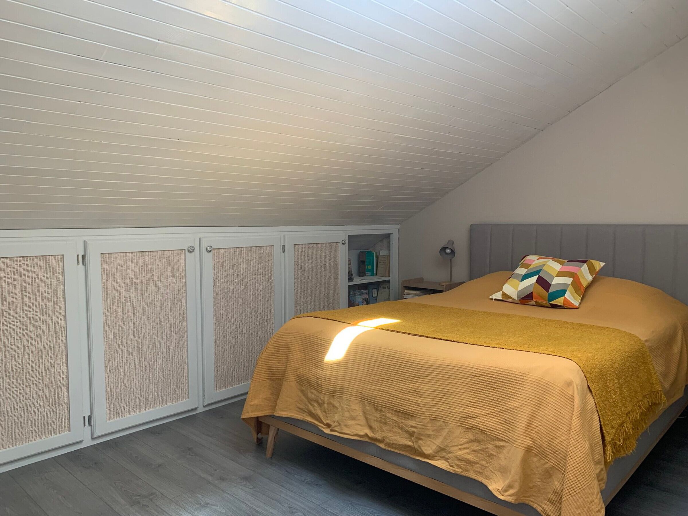 A bedroom with a sloped ceiling, a bed with a textured bedspread, a colorful pillow, and a nightstand with a lamp.