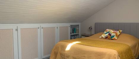 A bedroom with a sloped ceiling, a bed with a textured bedspread, a colorful pillow, and a nightstand with a lamp.