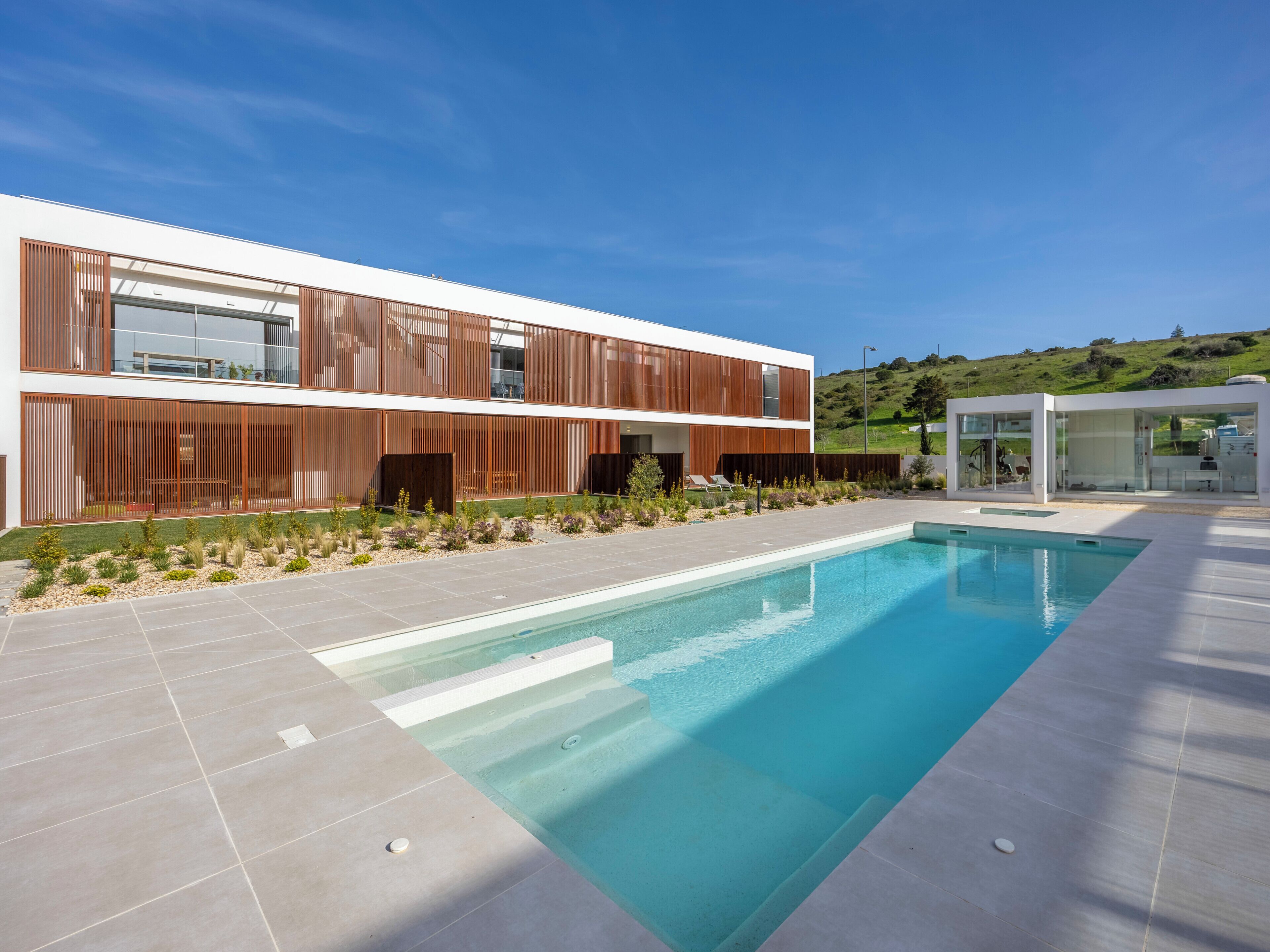 A modern building with a swimming pool, outdoor seating, and a view of a hill.