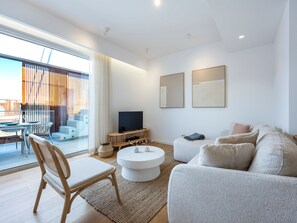 Ein modernes Wohnzimmer mit Sofa, Couchtisch und Fernseher. Ein Balkon mit Glastür und Blick ins Freie ist ebenfalls vorhanden.