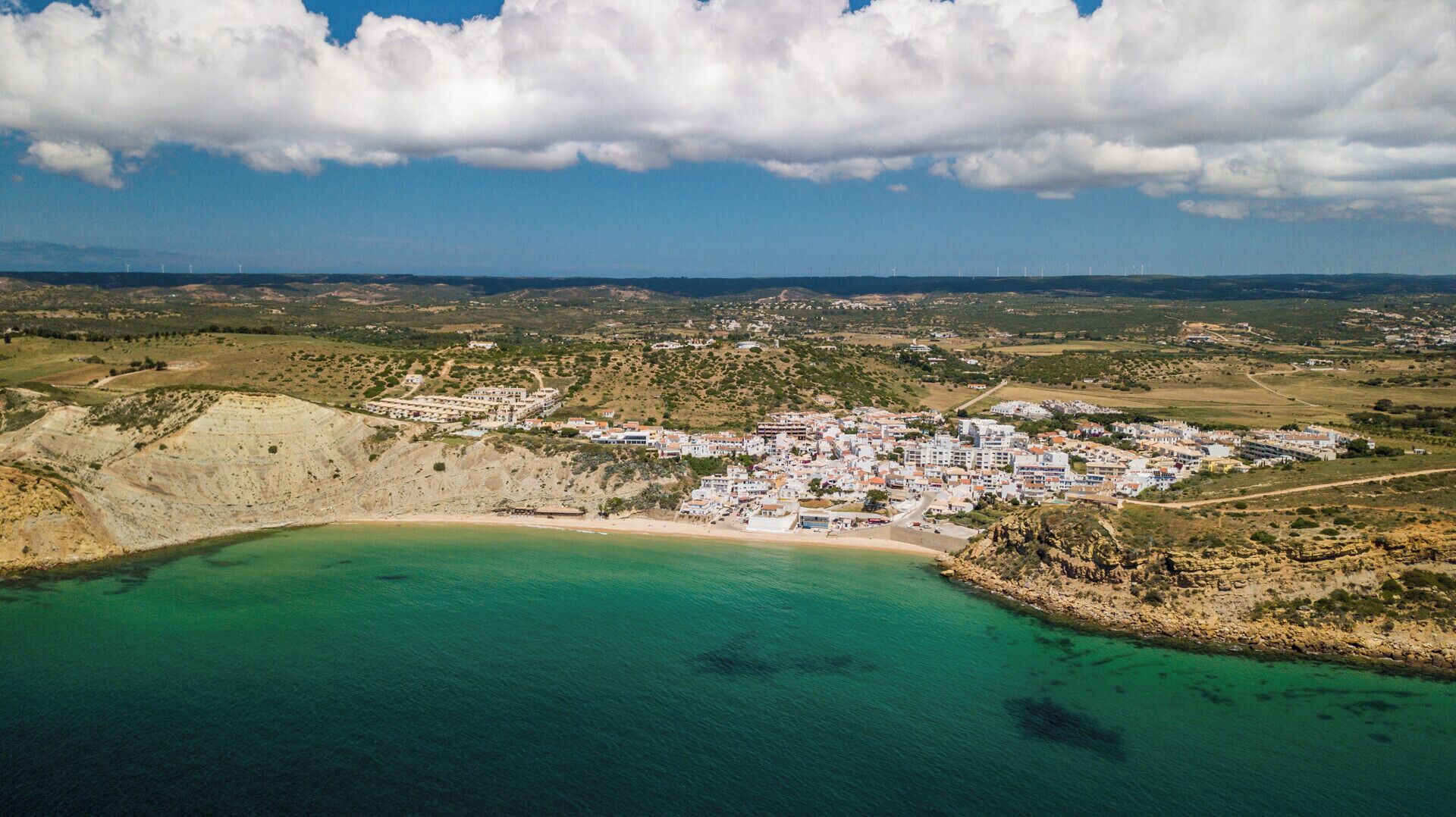 An aerial view of a coastal town with a beach, buildings, and wind turbines in the distance.
