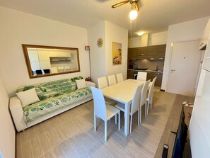 A compact living space with a dining area, kitchenette, and a sofa with a patterned cover. - Sea-view Apartment With Lovely, Furnished Terrace (Caorle)